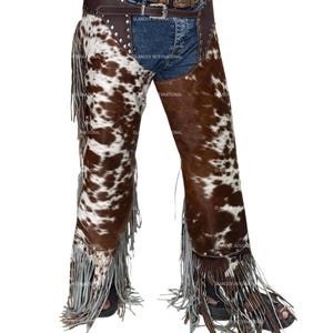 May include: Cowhide leather chaps with a brown and white spotted pattern. The chaps have a brown leather belt and fringe along the sides and bottom. The chaps are worn over blue jeans.