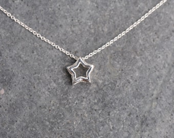 Puede incluir: Un delicado collar de plata con un colgante en forma de estrella hueca. La estrella es un diseño simple de cinco puntas, suspendido de una fina cadena. El collar se muestra sobre un fondo gris texturizado, resaltando el estilo minimalista de la joya.
