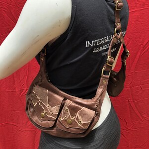 May include: A brown leather crossbody bag with multiple pockets and decorative stitching. The bag has a strap that goes over the shoulder and across the chest. The bag is decorated with metal studs and has a distressed look.