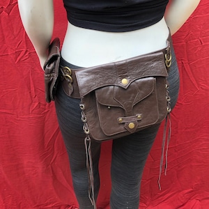 May include: Brown leather belt with a large pouch and two smaller pouches. The belt has decorative metal accents and buckles. The belt is worn on a mannequin.