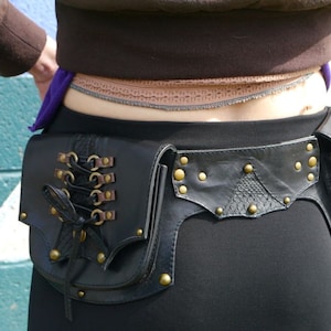 Pode incluir: Um cinto de couro preto com uma bolsa decorativa e um bolso com zíper. O cinto tem detalhes metálicos dourados e tachas.