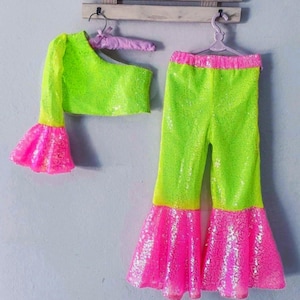 May include: A neon green sequined one-shoulder top with a long sleeve and flared cuff, paired with matching flared pants. The pants and sleeve cuffs are accented with bright pink sequins. The outfit is displayed on hangers against a light gray wall.