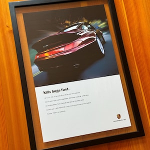 May include: Framed print featuring a maroon Porsche sports car speeding on a road. The print includes the text "Kills bugs fast" and other details about the car. The frame is black and the background is a wooden surface.
