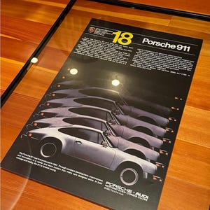 May include: A black and white framed poster with a black background and yellow text. The poster features a line of five silver Porsche 911 cars. The text on the poster reads "18 Porsche 911" and "Porsche + Audi Nothing Even Comes Close".