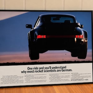 May include: A framed vintage Porsche advertisement poster featuring a rear view of a black sports car in mid-air against a twilight sky. The text reads: One ride and you'll understand why most rocket scientists are German, followed by descriptive copy.