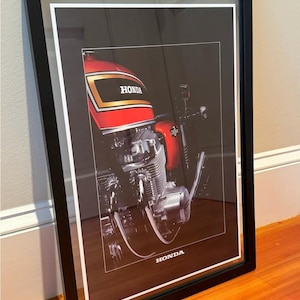 May include: Framed print of a classic Honda motorcycle. The motorcycle features a red fuel tank with gold accents and the Honda logo. The image is set against a dark background and framed in black.