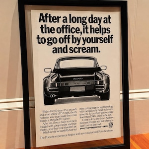 May include: Framed vintage Porsche advertisement. The black and white image features a black Porsche 911 Turbo from the rear. Text at the top reads, "After a long day at the office, it helps to go off by yourself and scream."