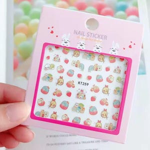 May include: A pink nail sticker sheet with a rectangular window displaying various cartoon characters, strawberries, and small houses. The sheet has the text "NAIL STICKER" and "KT259". The background is a blurred mix of colors.