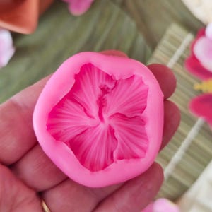 May include: A vibrant pink silicone flower-shaped mould. The mould features detailed petal impressions and a central pistil design. The mould is held in a hand, suggesting its size and use for crafting or baking.
