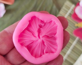 May include: A bright pink silicone mold in the shape of a flower. The mold has detailed petal impressions and a central pistil design. The mold is held in a hand, suggesting its size and use for crafting or baking.