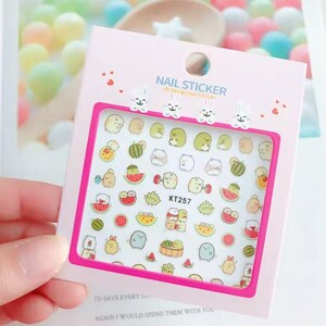 May include: A sheet of nail stickers featuring cartoon characters, watermelons, and other cute designs. The pink packaging reads "NAIL STICKER" and includes the text "KT257". The stickers are held by a hand.