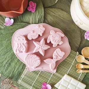 May include: A pink silicone mould with sea creature shapes: jellyfish, seahorse, starfish, turtle, seashell, and mermaid tail. The mould sits on a bamboo mat, with wooden spoons and pink flowers. The theme is ocean-themed baking, with measurements in cm.