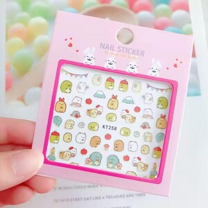 May include: A pink nail sticker sheet with a variety of colorful cartoon characters and objects. The sheet includes the text "NAIL STICKER" and the product code "KT258". The sheet is held in a person's hand.