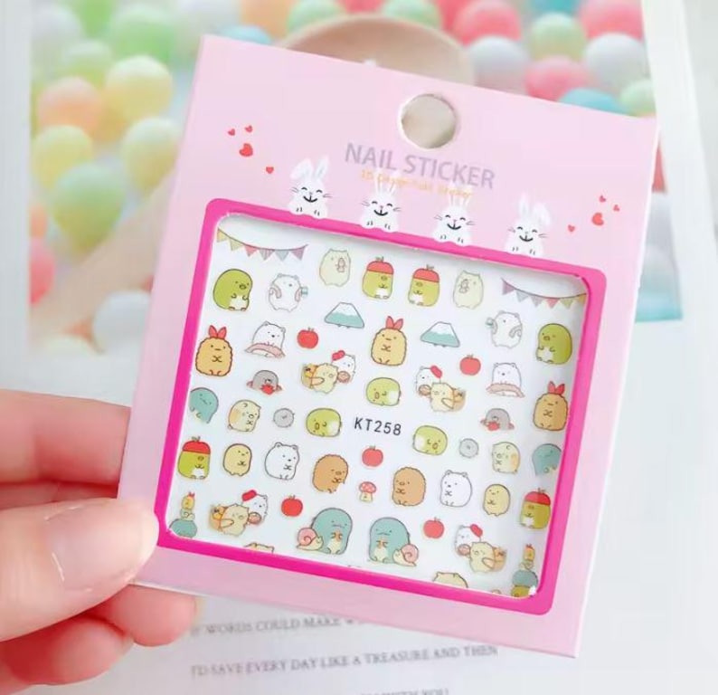 May include: A pink nail sticker sheet with a variety of colorful cartoon characters and objects. The sheet includes the text "NAIL STICKER" and the product code "KT258". The sheet is held in a person's hand.