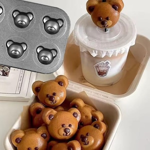 Könnte beinhalten: Ein Set brauner, bärenförmiger Cake Pops in einem weißen Papierbehälter. Die Cake Pops sind in einem Kreis angeordnet, wobei die Bärenköpfe nach außen zeigen. Ein einzelner, bärenförmiger Cake Pop auf einem Stiel befindet sich in einem durchsichtigen Plastikbecher mit weißem Deckel. Die Cake Pops werden in einer Metallbackform mit sechs bärenförmigen Formen hergestellt.