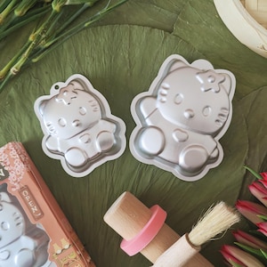 May include: Two silver-coloured, Hello Kitty-shaped cake tins. A wooden rolling pin with a pink silicone ring and a pastry brush are also visible. The tins are on a green surface, with a partially visible box that says "OPEN".