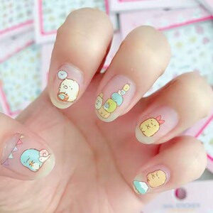 May include: A hand with clear nail polish decorated with colorful cartoon character nail art. The designs include a white bear with a blue cloud, a stack of colorful characters, and a yellow shrimp with a red bow.