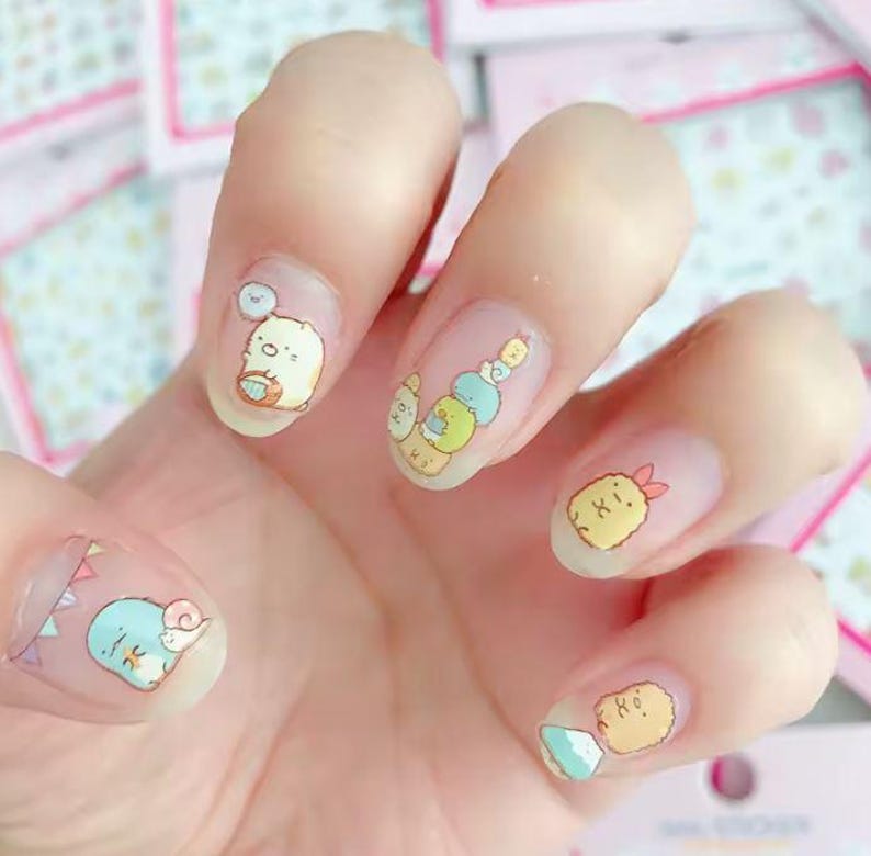 May include: A hand with clear nail polish decorated with colorful cartoon character nail art. The designs include a white bear with a blue cloud, a stack of colorful characters, and a yellow shrimp with a red bow.