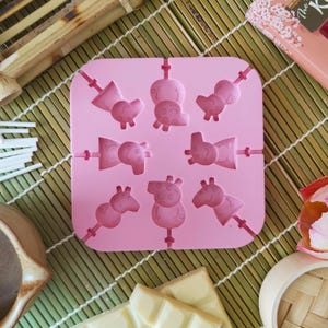 May include: A pink silicone mold for making character-shaped lollipops. The mold features various cartoon pig designs on lollipop sticks. The mold is square and sits on a bamboo mat, with white chocolate and other items in the background.