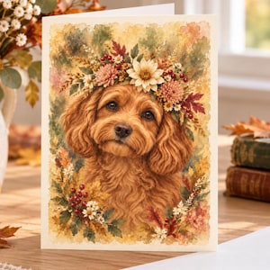 May include: A rectangular greeting card featuring a watercolor illustration of a fluffy golden-brown dog wearing a floral crown made of autumn leaves, white daisies, pink chrysanthemums, and red berries against a soft, textured background.