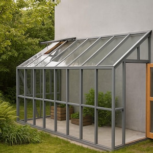 Puede incluir: Un pequeño invernadero moderno con techo y paredes de cristal y una puerta de madera. La estructura es gris y está unida a una pared gris claro. La puerta está abierta y hay plantas dentro. El invernadero está rodeado de hierba verde y árboles.