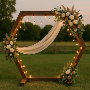 Puede incluir: Un arco de boda hexagonal de madera decorado con rosas blancas y melocotón, vegetación y luces de cadena. Una tela blanca transparente cuelga de la parte superior. El arco está sobre un campo de hierba con un fondo borroso.