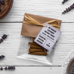 Op de afbeelding: Een kleine doorzichtige plastic zak gevuld met zes bruine wierookkegels. De zak is vastgebonden met een bruin touwtje en heeft een wit label met de tekst "All-Natural Lavender Incense Cones" en "Lavande - Cones d'Encens". Het label vermeldt ook "6 Cones".