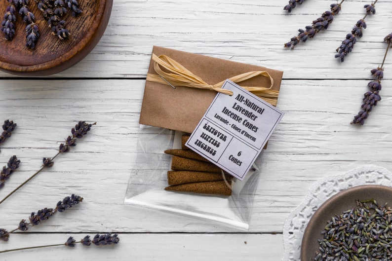 Op de afbeelding: Een kleine doorzichtige plastic zak gevuld met zes bruine wierookkegels. De zak is vastgebonden met een bruin touwtje en heeft een wit label met de tekst "All-Natural Lavender Incense Cones" en "Lavande - Cones d'Encens". Het label vermeldt ook "6 Cones".