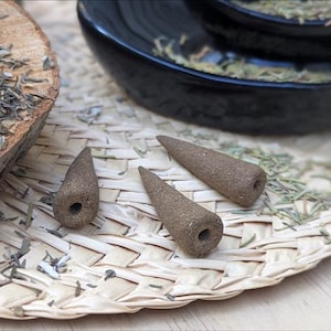 Könnte beinhalten: Drei braune, kegelförmige Räucherkegel mit strukturierter Oberfläche und einem kleinen Loch an der Basis. Die Räucherkegel sind auf einer geflochtenen Unterlage neben einer Holzscheibe und einer schwarzen Schale mit getrockneten Kräutern angeordnet.