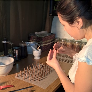 Pictured is the owner of Leftover Hippies Incense rolling cones by hand. The surface in front of Hazel has a mixing bowl, grinder, mortar and pestle, and many jars of finished incense sticks on it. There are several lines of newly completed cones in front of her hands, which are at work.