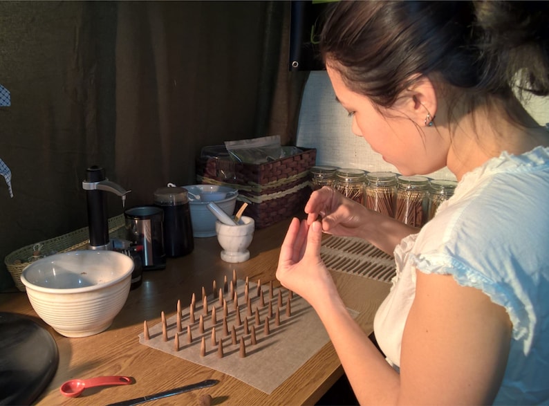 Pictured is the owner of Leftover Hippies Incense rolling cones by hand. The surface in front of Hazel has a mixing bowl, grinder, mortar and pestle, and many jars of finished incense sticks on it. There are several lines of newly completed cones in front of her hands, which are at work.