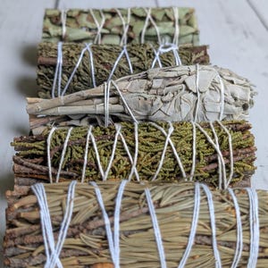 Puede incluir: Una colección de cinco manojos cilíndricos de hierbas secas y agujas de hoja perenne, cada uno envuelto firmemente con hilo de algodón blanco. Los manojos presentan texturas y tonos variados de verde, marrón y blanco, dispuestos en fila.