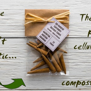 Op de afbeelding: Een doorzichtige plastic zak gevuld met 12 bruine wierookkegels. De zak is vastgebonden met een bruin lint en een label met de tekst "All-Natural Lavender Incense Cones" en "12 Cones". De tekst "These bags aren't plastic... They're plant cellulose! 100% compostable" staat op een witte houten achtergrond.