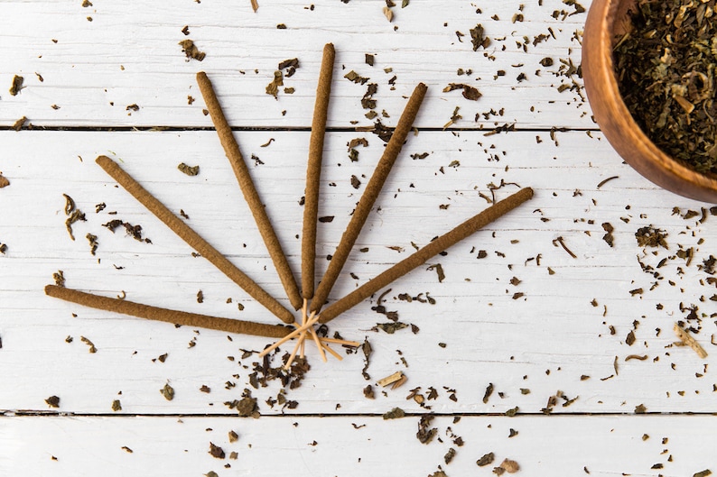 May include: A set of brown incense sticks arranged in a fan shape on a white wooden surface. The sticks are surrounded by dried herbs.