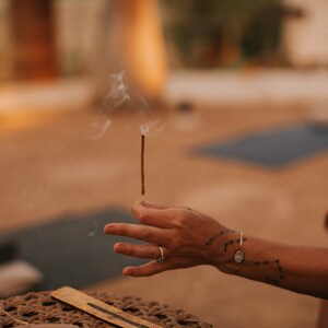 An incense stick with a glowing, burning tip emitting a small wisp of smoke. The stick is held vertically by a tattooed hand, showcasing its textured surface and the ember at the end.