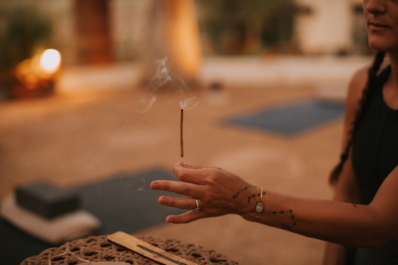 An incense stick with a glowing, burning tip emitting a small wisp of smoke. The stick is held vertically by a tattooed hand, showcasing its textured surface and the ember at the end.