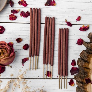 Peut inclure: Un ensemble de trois paquets de b&acirc;tonnets d'encens bruns sur une surface en bois blanc avec des p&eacute;tales de rose s&eacute;ch&eacute;s &eacute;parpill&eacute;s autour.