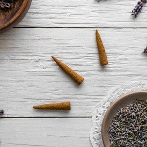 3 hand-rolled natural Lavender incense cones on a white wooden surface with dried lavender flowers scattered around.