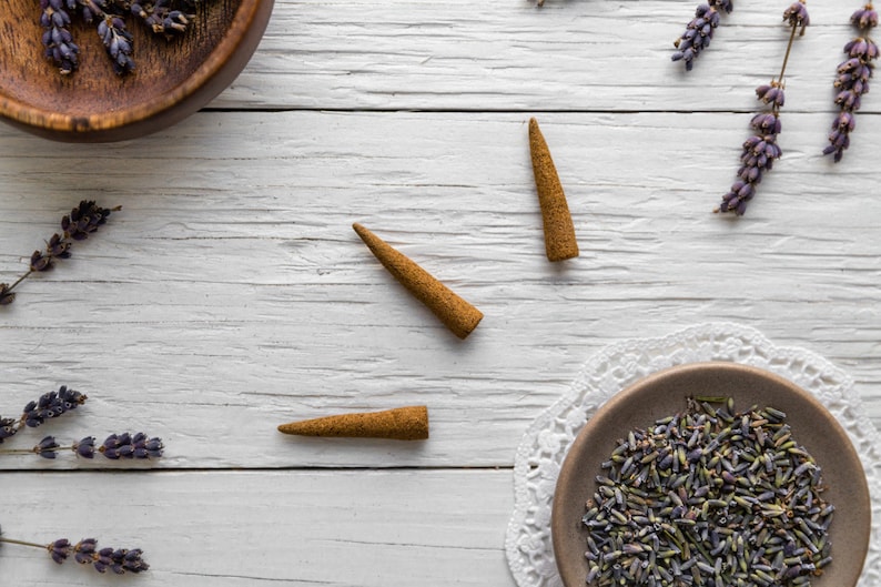 3 hand-rolled natural Lavender incense cones on a white wooden surface with dried lavender flowers scattered around.