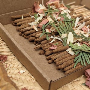 Bulk box of 60 Cedar Incense Sticks topped with dried green cedar sprigs and scattered cedar wood chips.