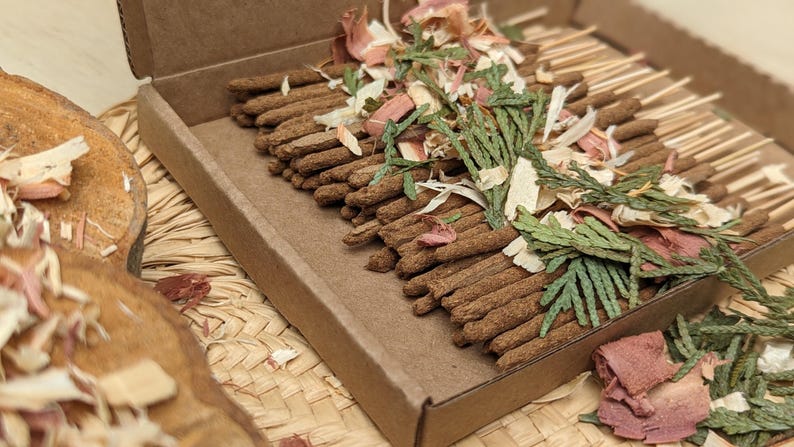 Bulk box of 60 Cedar Incense Sticks topped with dried green cedar sprigs and scattered cedar wood chips.