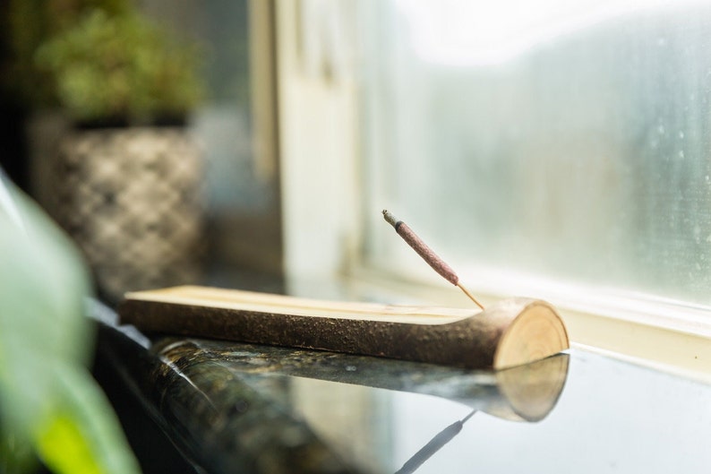 A natural wood incense holder with a rough, bark-covered exterior and a smooth, carved interior channel. A lit incense stick is inserted into the holder, with a small amount of grey ash visible at the top of the stick.