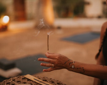 Pode incluir: Um incenso fino e castanho que emite um rasto de fumo branco. O incenso é segurado na vertical entre os dedos, posicionado sobre uma base circular entrançada que contém um suporte de madeira com vestígios de cinza na superfície.