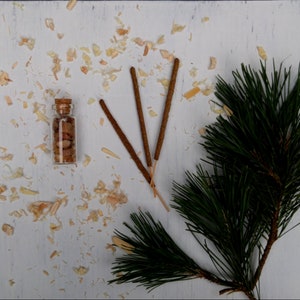 May include: A small glass bottle with a cork stopper filled with brown and white crystals, three brown incense sticks, and a green pine branch on a white wooden surface.