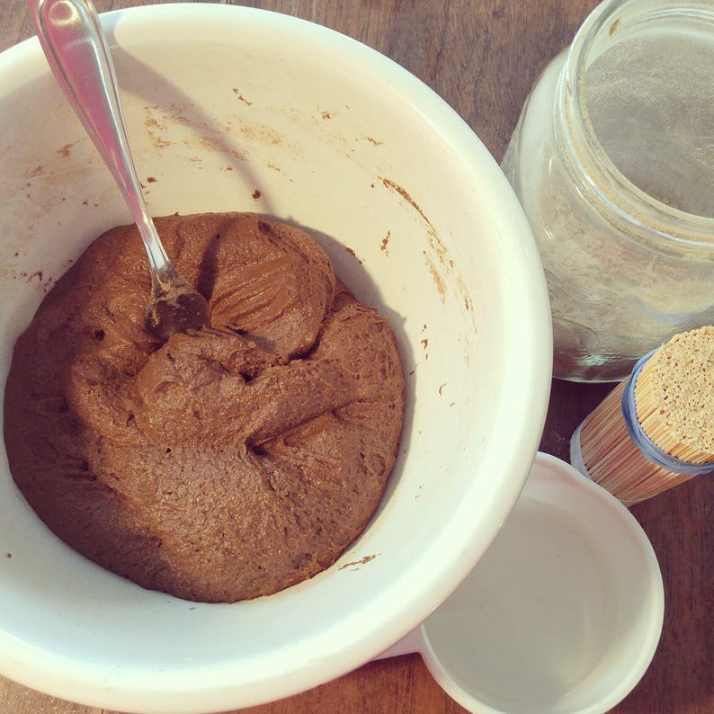 A bowl of incense dough in a mixing bowl with a fork sticking out of it. Beside the bowl are a measuring cup, a bundle of bamboo stick, and a jar of powdered acacia gum.