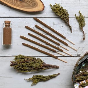 May include: A top-down view showcasing natural incense and related items. The image includes brown incense sticks, a small glass bottle with a cork, dried green sprigs, and a wooden slice, arranged on a white wooden surface, creating a natural aesthetic.