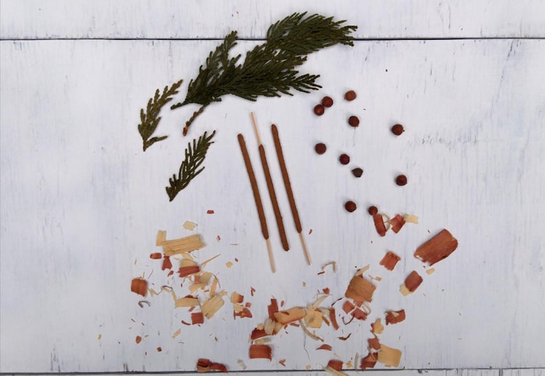 3 Cedar Incense Sticks arranged in a row, accompanied by several green cedar sprigs, small cedar berries, and scattered cedar wood shavings on a white wooden surface.