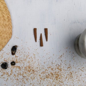 Puede incluir: Tres conos de incienso marrones sobre una superficie blanca con un sombrero de paja tejido y un mortero y un pilón blancos en el fondo.