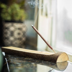Peut inclure: Un porte-encens en bois avec une finition en &eacute;corce naturelle et un seul b&acirc;ton d'encens br&ucirc;lant, cr&eacute;ant une tra&icirc;n&eacute;e de fum&eacute;e. Le porte-encens est pos&eacute; sur un comptoir sombre.