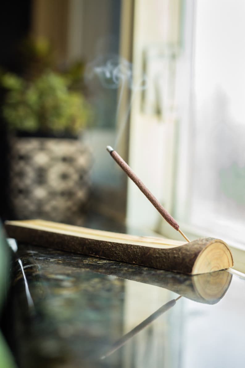 Peut inclure: Un porte-encens en bois avec une finition en &eacute;corce naturelle et un seul b&acirc;ton d'encens br&ucirc;lant, cr&eacute;ant une tra&icirc;n&eacute;e de fum&eacute;e. Le porte-encens est pos&eacute; sur un comptoir sombre.